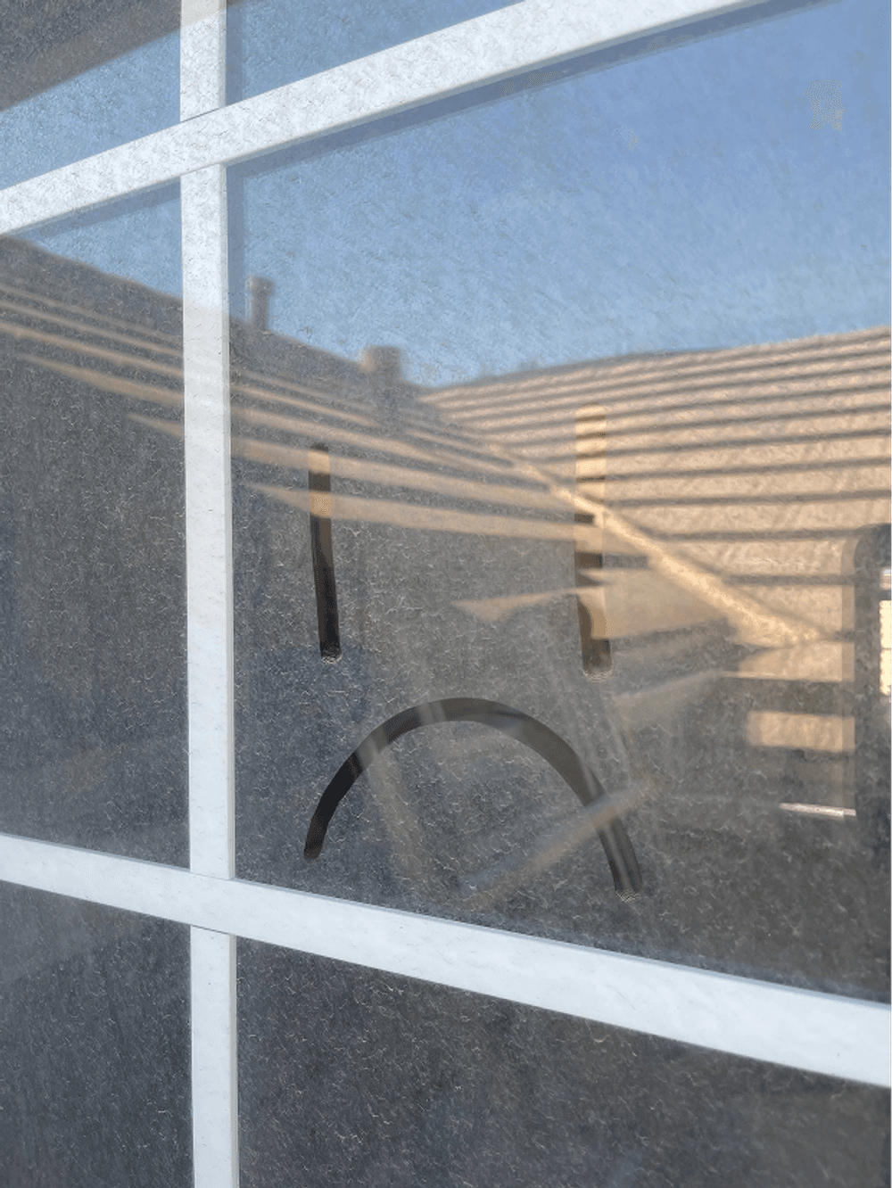 Smudged glass with a sad face drawn on it, reflecting sunlight and a rooftop background.