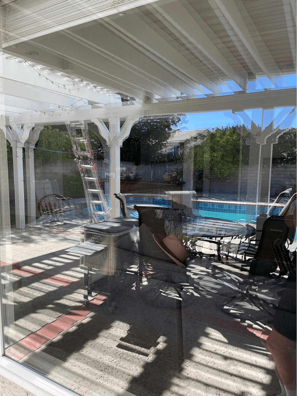 Backyard patio with pool, chairs, and ladder under covered area, illuminated by sunlight.