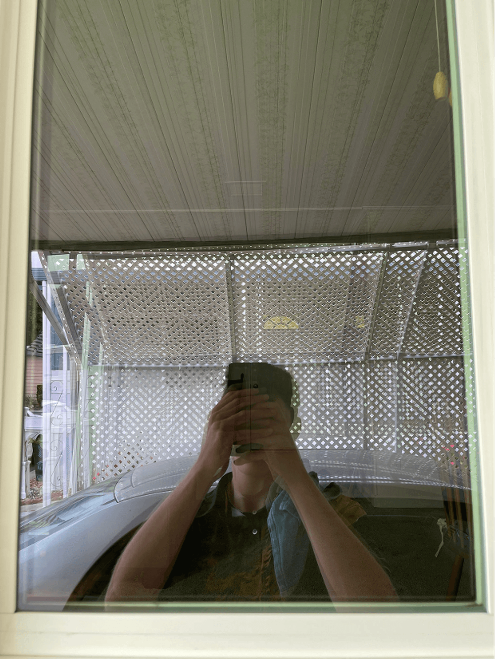 Reflection of a person holding a phone in a window, with a car visible outside.
